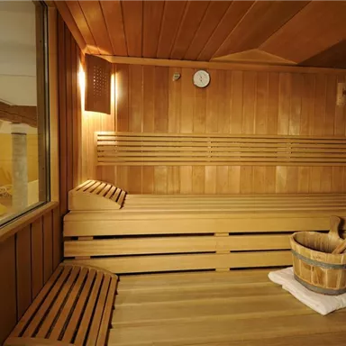 A cozy wellness area with a wooden bench and a wooden bucket. The walls are made of light wood and there is a large window cutout.