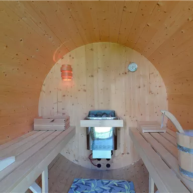 Ein gemütlicher Saunaraum aus Holz mit Bänken und einer ruhigen Atmosphäre. In der Mitte befindet sich ein Ofen und ein Eimer aus Holz.