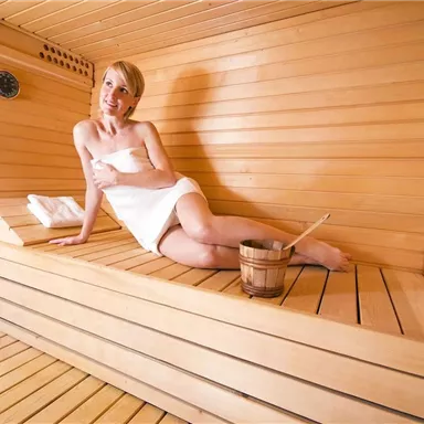 Eine entspannte Person sitzt in einer Sauna aus Holz. Sie trägt ein Handtuch und genießt die Ruhe und Wärme.