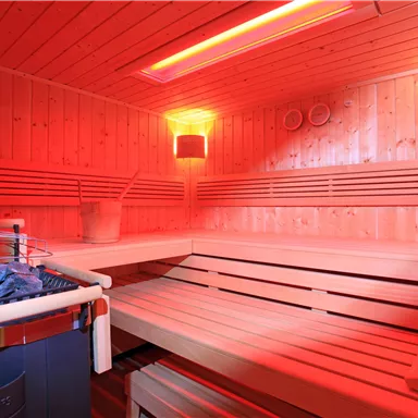 A modern sauna with wood paneling and red light. In the foreground, stones and a wooden container can be seen.