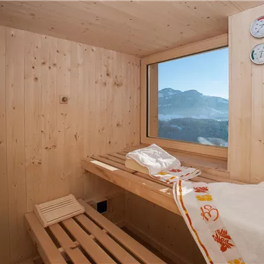 Ein gemütlicher Saunaraum mit Holzverkleidung und einer großen Fensteransicht in die Berge. Auf den Bänken liegen Handtücher bereit für die Entspannung.