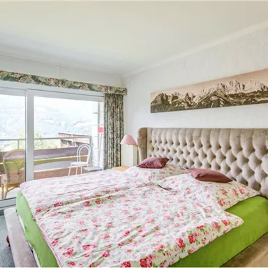 Ein gemütliches Schlafzimmer mit einem großen Bett und floralen Bettwäsche. Durch die Fenster hat man einen schönen Blick auf die Berge und die Terrasse.
