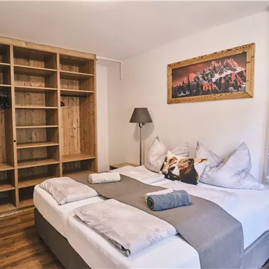 A cozy bedroom with two beds, wooden shelves, and a stylish lamp. On the wall hangs a picture of a mountain landscape.