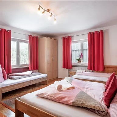 A cozy bedroom with two beds and bright red curtains. The room has large windows and a warm wooden floor.