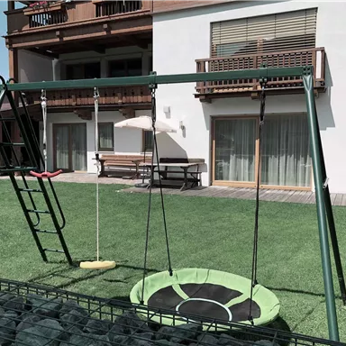 A playground with a swing and a seesaw in a garden. In the background, a house and a terrace with windows can be seen.