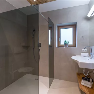 A modern bathroom with a glass shower and a white sink. Light grey walls and wooden shelves give the space a warm atmosphere.
