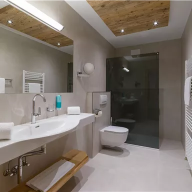 A modern bathroom with a large sink and a glass shower. The walls are light and the ceiling has wooden elements.