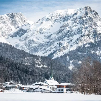 A picturesque mountain landscape with snow-covered peaks and a small village in the foreground. The sky is clear and the surroundings are wintery calm.