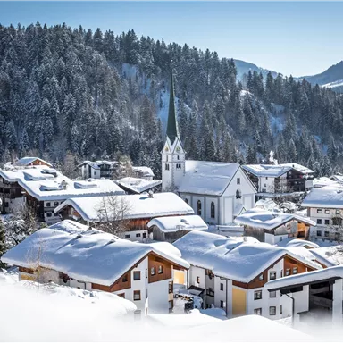 A picturesque village in the snow with snow-covered houses. In the background, there is a church and forested mountains visible.