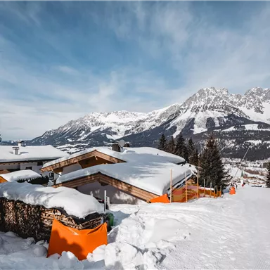 Eine verschneite Landschaft mit Hütten und hohen Bergen im Hintergrund. Der klare Himmel verstärkt die winterliche Stimmung.