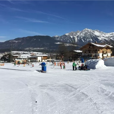 A snow-covered landscape with mountains in the background. In the foreground, there are people skiing and playing in the snow.