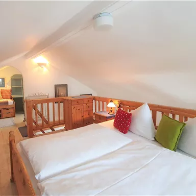 A cozy attic room with a large bed and wooden furniture. There is warm lighting and colorful cushions for added comfort.