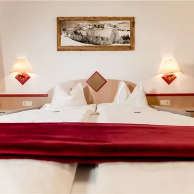 Das Bild zeigt ein gemütliches Hotelzimmer mit zwei Betten und roten Akzenten. An den Wänden hängen dekorative Lampen und ein Bild von einer Berglandschaft.