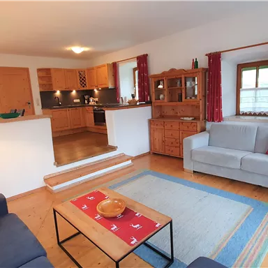A cozy living room with wooden flooring and a modern kitchen. The furniture is simple and inviting, with red curtains and a rug in soft colors.