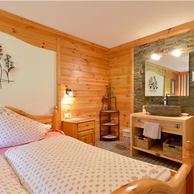 Ein gemütliches Schlafzimmer aus Holz mit einem Doppelbett und dekorativen Kissen. Der Raum bietet auch ein Badezimmer mit modernen Accessoires.