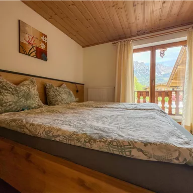 Ein gemütliches Schlafzimmer mit einem Doppelbett und zwei Kissen. Die großen Fenster bieten einen Blick auf die Berge und viel Tageslicht.