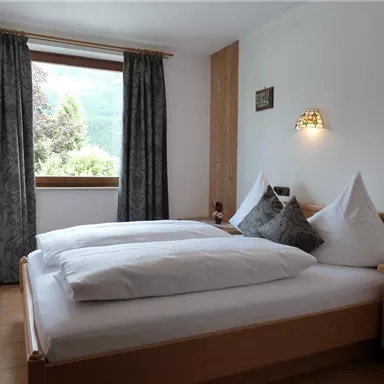 A cozy bedroom with a double bed and two pillows. Large windows decorated with curtains offer a view of nature outside.