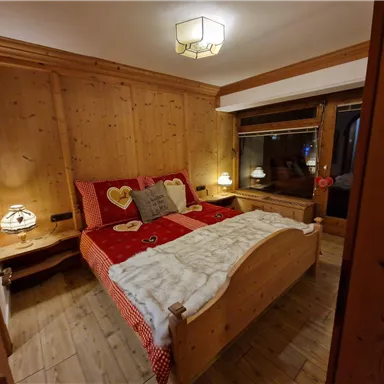 A cozy bedroom with wooden walls and a large bed. The bedding is red with heart patterns and there is soft lighting.