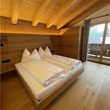 A cozy bedroom with a large double bed and wooden paneling. Bright walls and a balcony window create an inviting atmosphere.