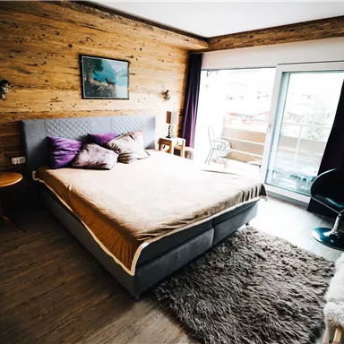 A cozy bedroom with a large bed and wooden paneling. There is a balcony and modern furniture.