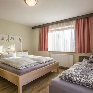 Ein modernes Schlafzimmer mit einem Doppelbett und Einzelbett. Helle Fenster und gemütliche Dekoration sorgen für eine einladende Atmosphäre.