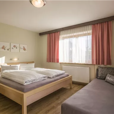 Ein helles Schlafzimmer mit einem Doppelbett und einem gemütlichen Sofa. Große Fenster lassen viel Tageslicht herein und verleihen dem Raum eine einladende Atmosphäre.