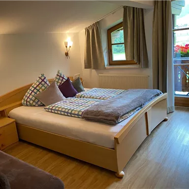 Ein gemütliches Schlafzimmer mit einem großen Bett und modernen Holzmöbeln. Große Fenster lassen viel Tageslicht herein und bieten einen Blick auf einen Balkon mit Blumen.