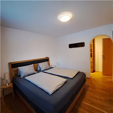 A bright bedroom with a large bed and wooden floor. The walls are white, and there is an open access to another room.