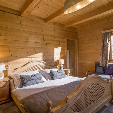 Ein gemütliches Schlafzimmer mit Holzverkleidung und großen Fenstern. Ein Bett mit weißen und grauen Kissen sowie ein bequemer Sessel laden zum Entspannen ein.