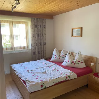 Ein gemütliches Schlafzimmer mit einem Doppelbett und bunten Kissen. Der Raum hat Holzdetails und große Fenster, die viel Tageslicht hereinlassen.
