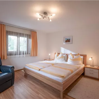 A cozy bedroom with a large bed and bright curtains. There is a comfortable armchair and a modern lamp.