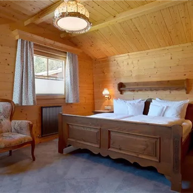 A cozy wooden room with a double bed and soft colors. There is a comfortable armchair and windows with curtains that let in gentle light.