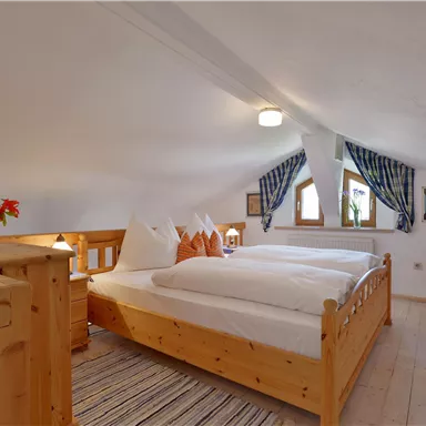 A cozy bedroom with wooden furniture and two beds. The windows are decorated with checked curtains and there are fresh flowers on the nightstand.