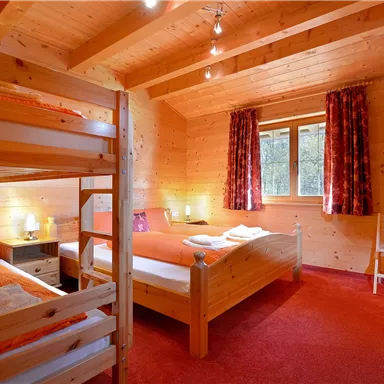 A cozy wooden room with two beds and a loft bed. The room features orange accents and large windows overlooking nature.