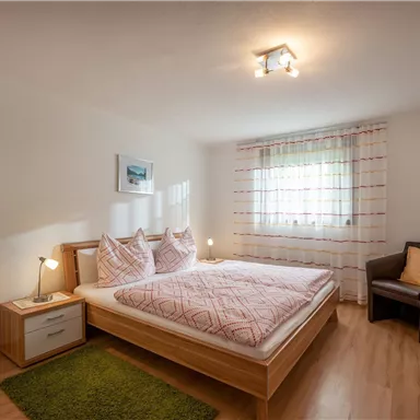 A cozy bedroom with a double bed and bright textiles. There is a window with curtains and a comfortable armchair next to it.