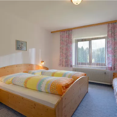 A bright bedroom with two beds and an additional sofa bed. Large windows let in plenty of daylight and the walls are decorated with floral curtains.