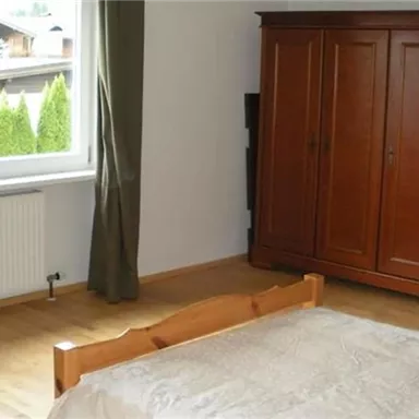Ein schlichtes Schlafzimmer mit einem Holzschrank und einem großen Fenster. Der Boden ist aus Holz und es gibt einen Bett mit einer einfachen Bettdecke.