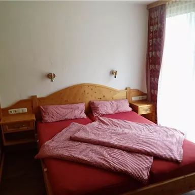 Ein gemütliches Schlafzimmer mit einem Doppelbett und roten Bettdecken. Das Zimmer hat Holzschränke und große Fenster, die viel Licht hereinlassen.