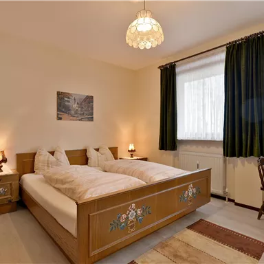 A cozy bedroom with two beds, wooden furniture, and a small table. Plenty of daylight comes through the windows, and the walls are painted in neutral colors.