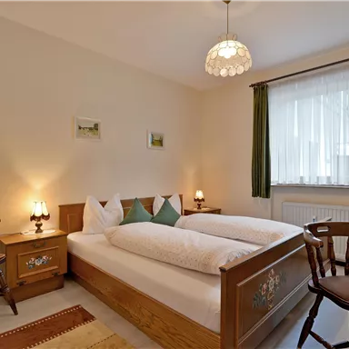 A cozy bedroom with a double bed and wooden chair. Large windows with curtains provide daylight and an inviting atmosphere.