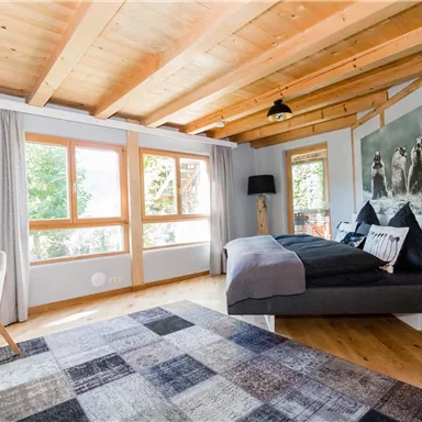 Ein gemütliches Schlafzimmer mit Holzbalkendecke und großen Fenstern. Es enthält ein modernes Bett, einen Schreibtisch und einen Teppich.