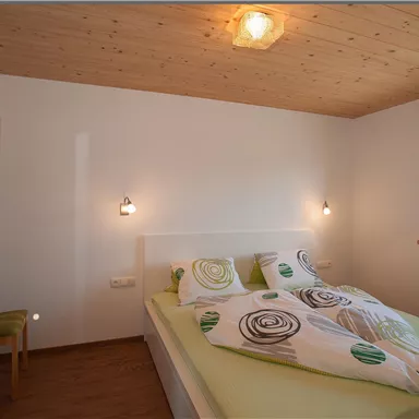 Ein gemütliches Schlafzimmer mit einem großen Bett und bunten Bettdecken. Die Wände sind hell und es gibt ein Fenster, das viel Licht hereinlässt.