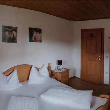 A cozy bedroom with a double bed and wooden furniture. Pictures hang on the walls, and the room has large windows with cheerful curtains.