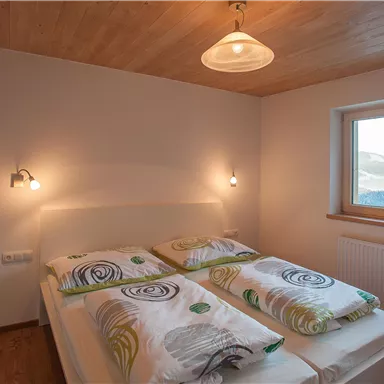 Ein modernes Schlafzimmer mit einem großen Fenster und Holzdecke. Zwei Betten sind mit farbigen Bettdecken dekoriert.