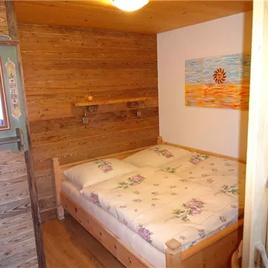 A cozy bedroom with a wooden frame bed and blooming pillows. The walls are made of wood, and on one wall hangs a colorful picture.