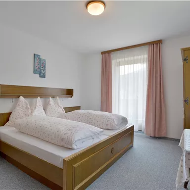 A simple bedroom with two single beds and wooden paneling. The room has large windows and a small table.