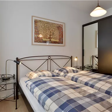 A cozy bedroom with a double bed and checkered bedspreads. A piece of art hangs on the wall, and a black wardrobe stands next to the bed.