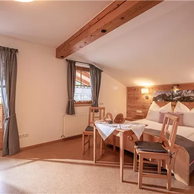 A cozy room with a double bed and wooden decor. The windows offer a view outside and there is a table with chairs.