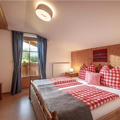 A cozy bedroom with a double bed, wooden panels, and checked cushions. Large window view with blue curtains and bright wooden furniture.