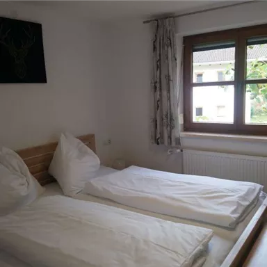 A cozy bedroom with two single beds and white bedspreads. A window overlooking the outdoors provides natural light.